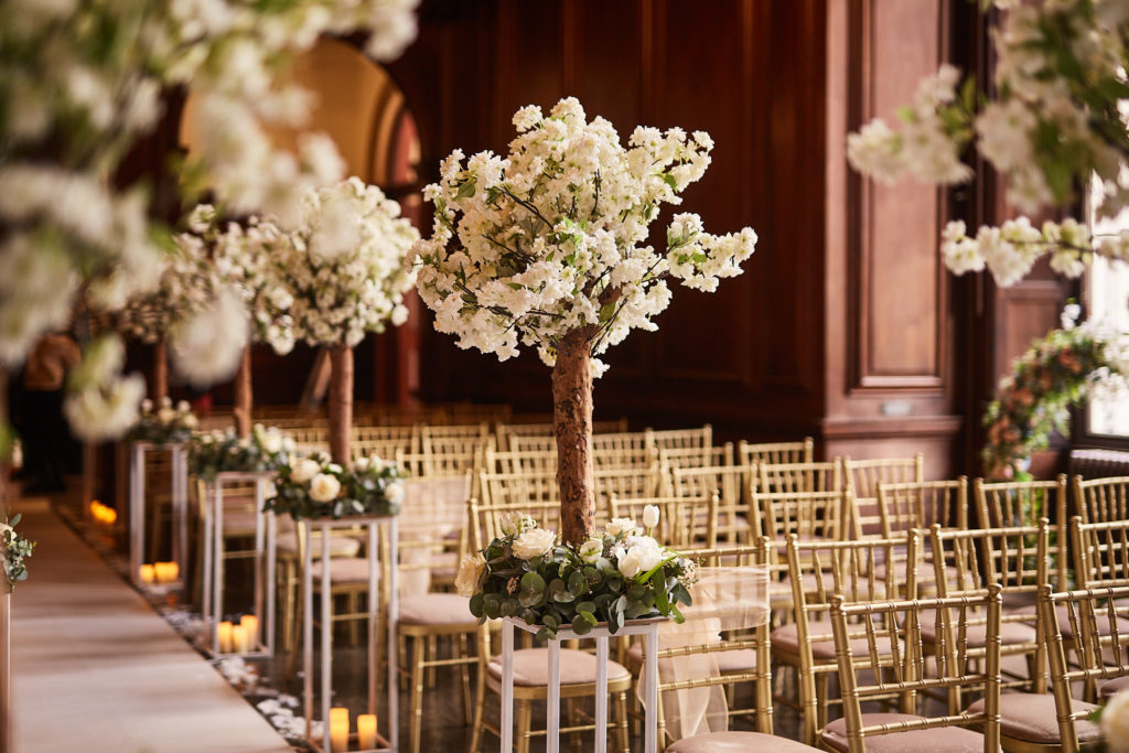 Ceremony room lined with white blossom trees and gold chairs at Addington Palace