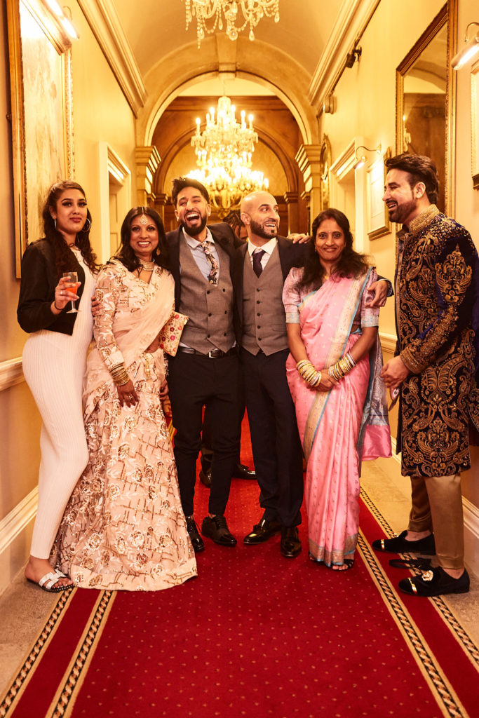 Group of friends in Asian wedding outfits and suits in a chandelier-lit corridor at Addington Palace