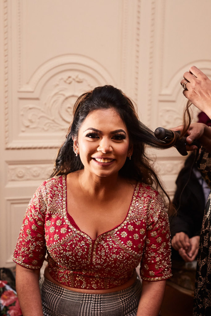 Bride smiling in a red and gold embroidered lehenga as her hair is styled