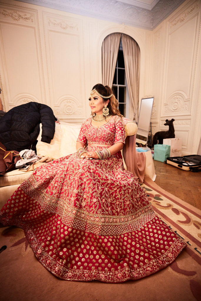 Bride in a red and gold lehenga sitting on a sofa with her skirt spread out