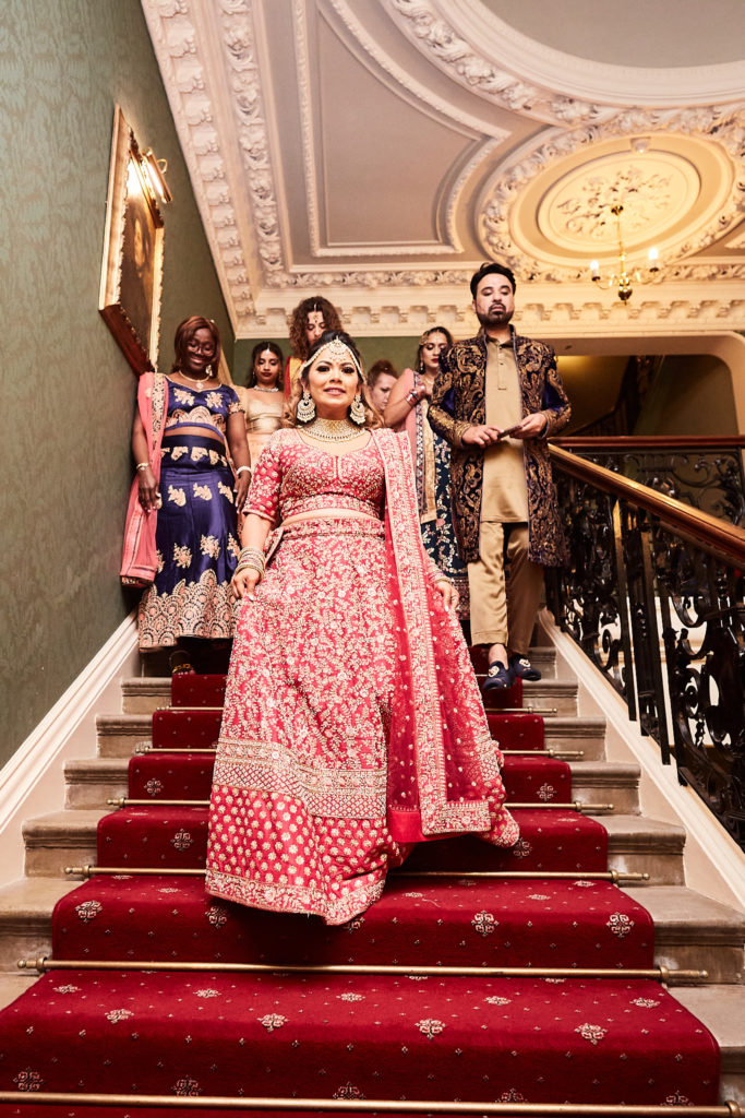 Bride in a red lehenga coming down the grand staircase at Addington Palace with her bridal party