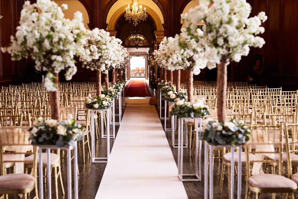 White aisle lined with blossom trees and gold chairs in the Great Hall at Addington Palace