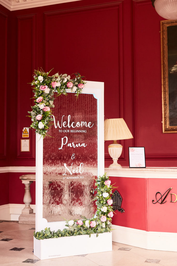 Mirrored welcome sign for Parna and Neil's wedding, dressed with flowers, in the red hallway at Addington Palace