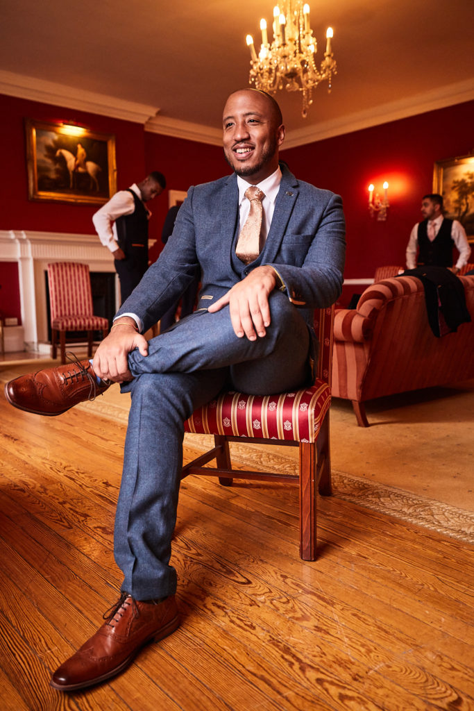 Groom in a blue tweed suit sitting with his legs crossed in a red drawing room at Addington Palace