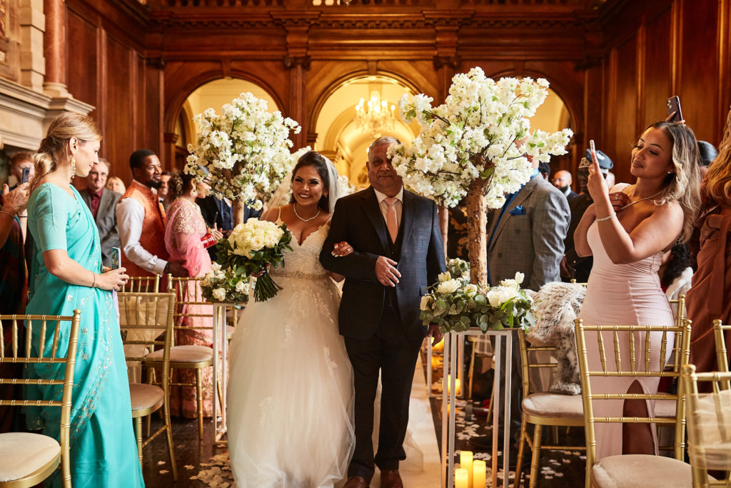 Bride walking down the aisle on the arm of an older man as guests take photos
