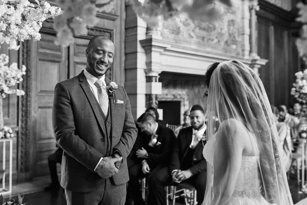 Black and white photo of the groom smiling at the bride as guests look on