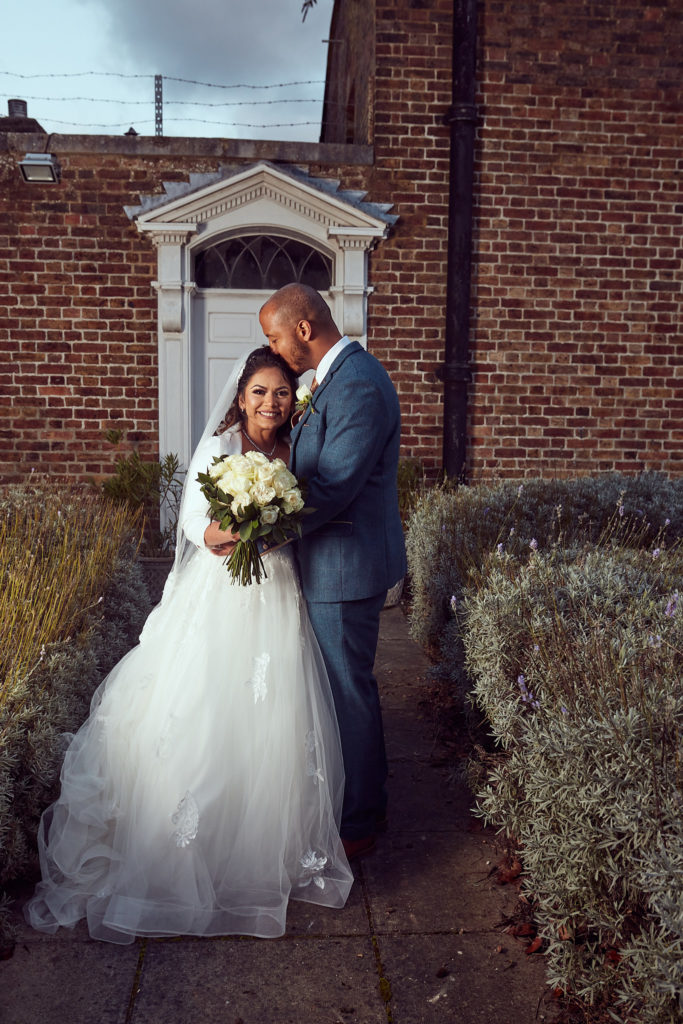 Groom kissing the bride's forehead beside a red-brick wall at Addington Palace