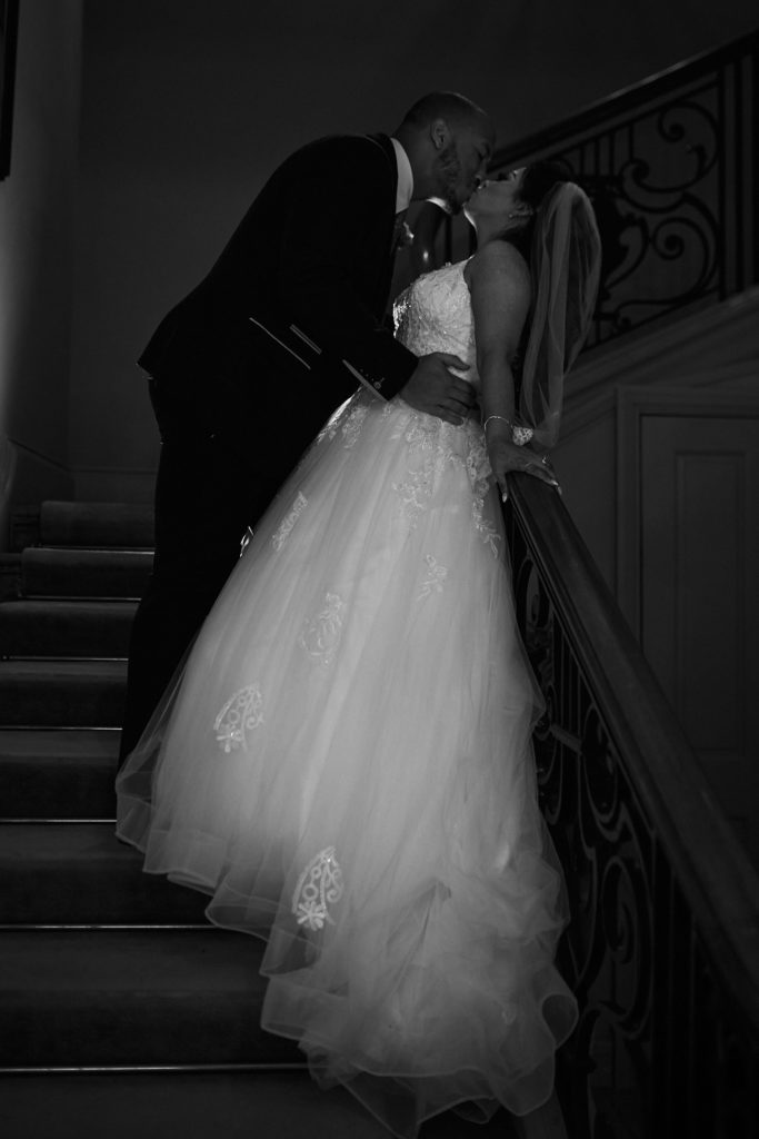 Black and white photo of the groom kissing the bride on the staircase at Addington Palace