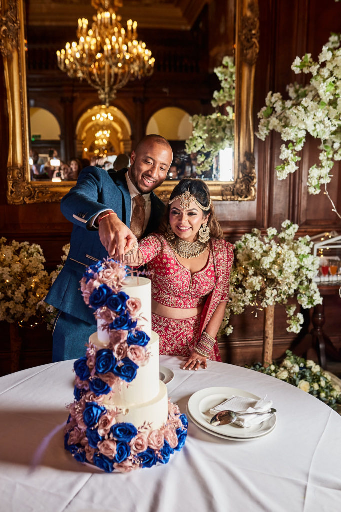 Bride in a red lehenga and the groom cutting their wedding cake under a chandelier