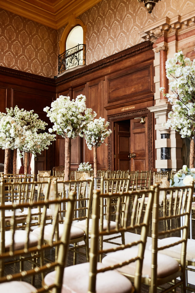Gold ceremony chairs and blossom trees beneath the wood-panelled gallery at Addington Palace