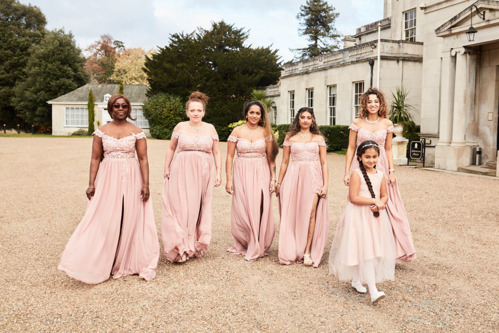 Bridesmaids in blush pink dresses and a flower girl walking across the drive at Addington Palace