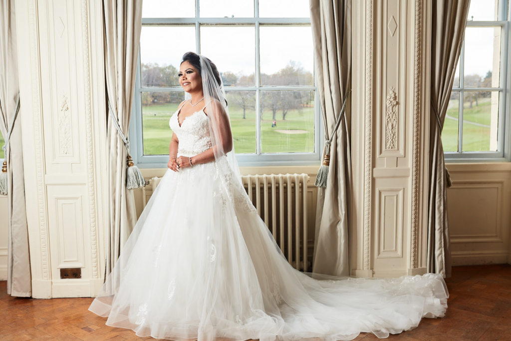 Bride in her wedding dress and veil standing between tall windows at Addington Palace