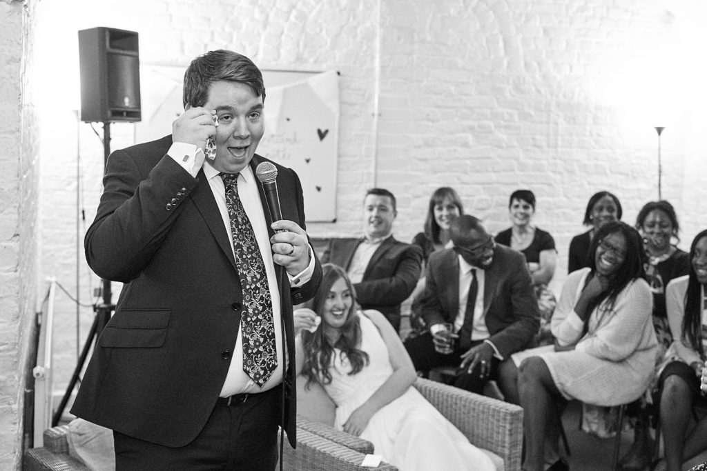 Black and white photo of the groom rubbing his eye as he gives his speech