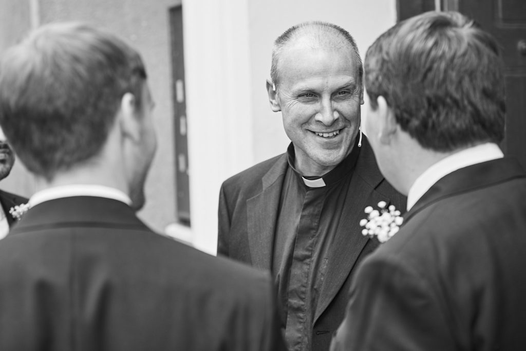Black and white photo of the vicar smiling as he chats with the groom