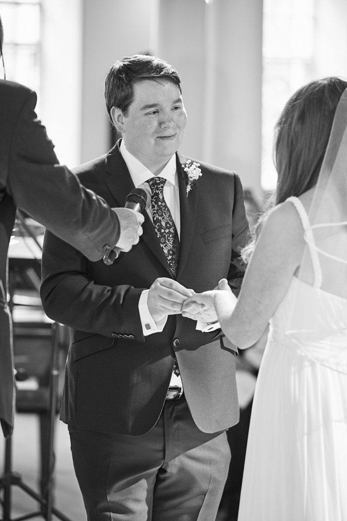 Black and white photo of the groom smiling as he holds the bride's hands during their vows