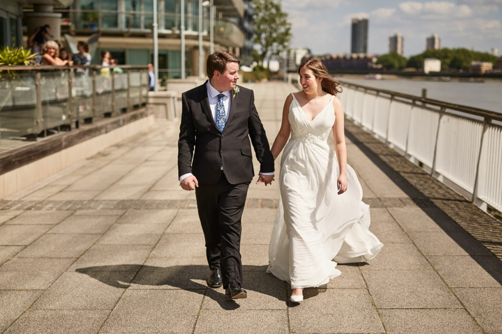 Bride and groom laughing as they walk hand in hand along the Thames path in London