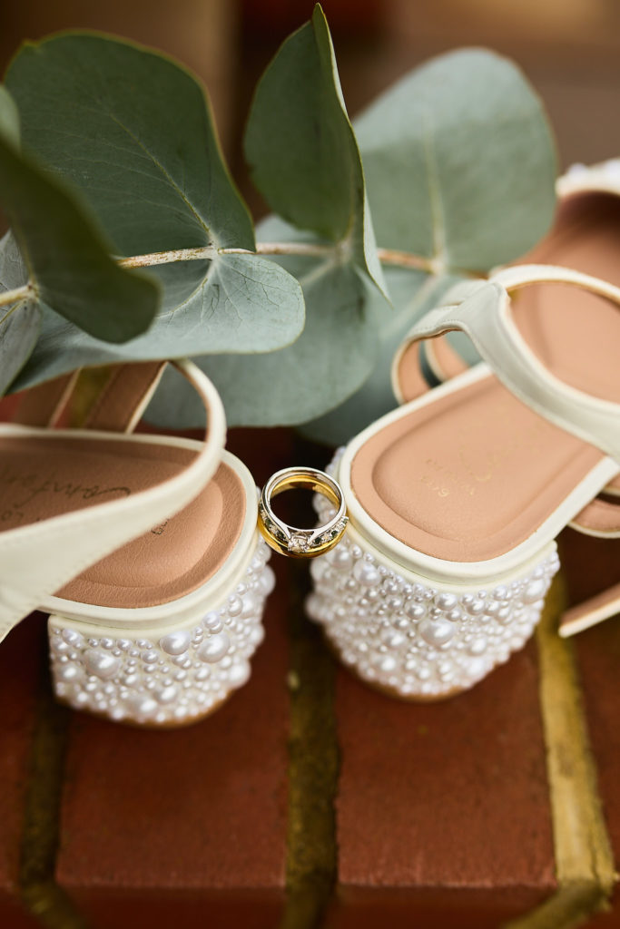 Pearl-heeled bridal shoes with a wedding ring balanced between them, eucalyptus leaves on red brick