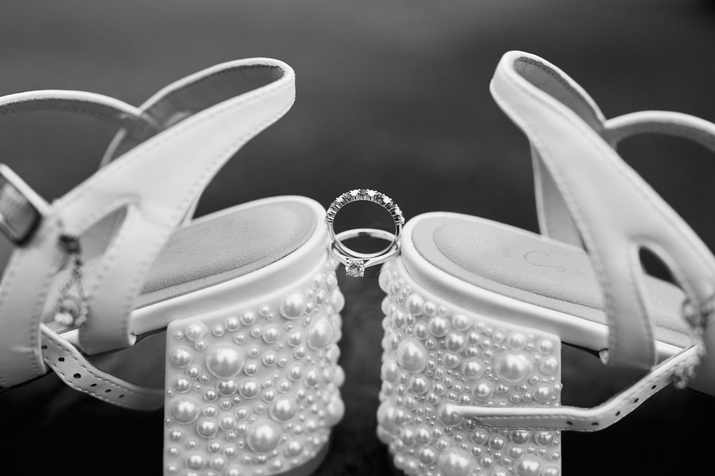 Black and white close-up of pearl block-heel bridal shoes with wedding rings between them