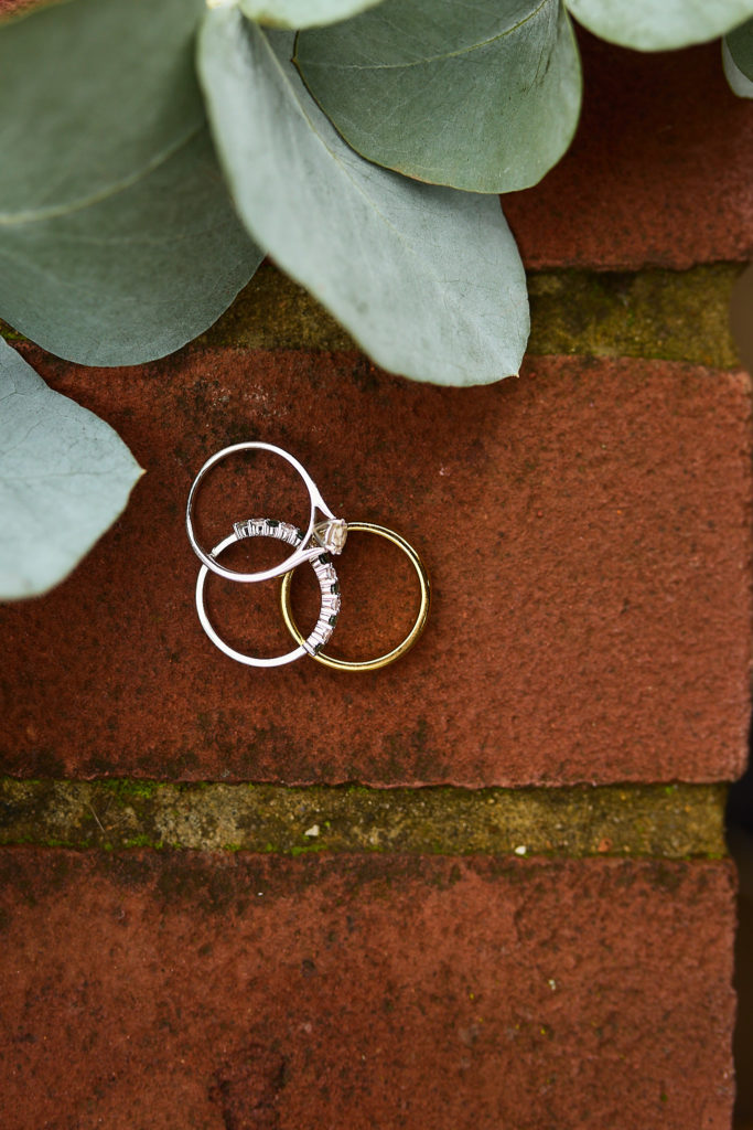 Wedding rings and an engagement ring on red brick beside eucalyptus leaves