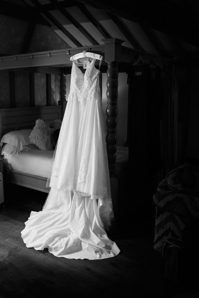 Black and white photo of a wedding dress hanging from a four-poster bed in an oak-beamed room