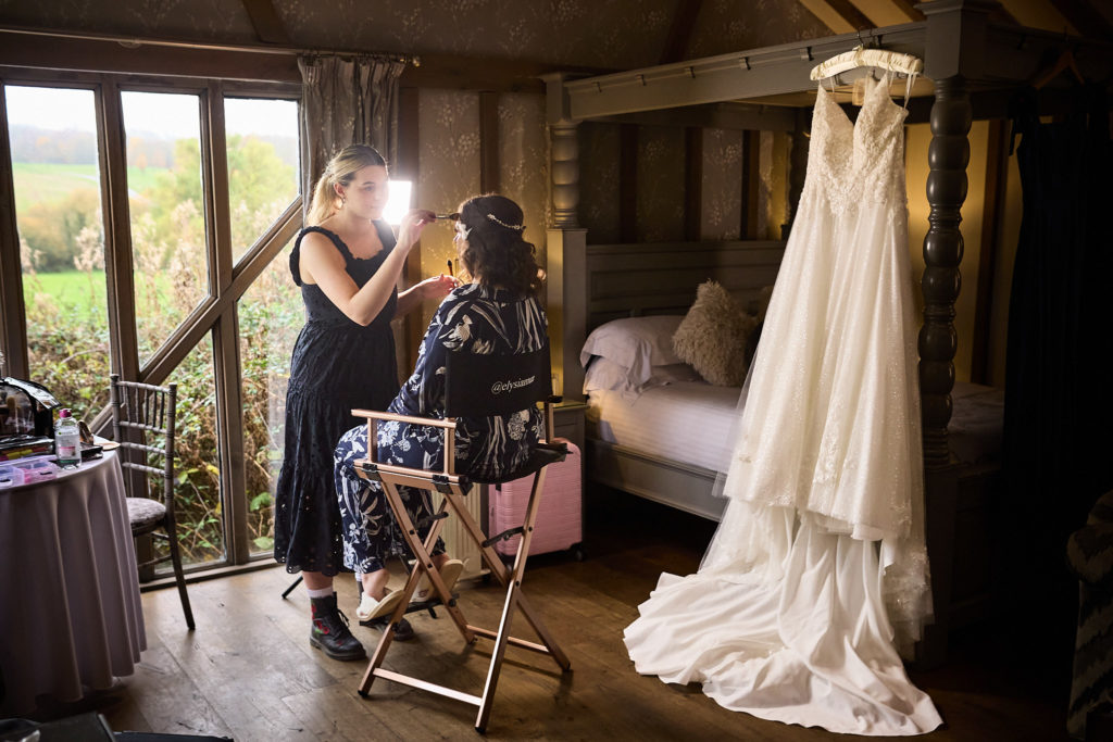 Bride having her make-up done by the window, her wedding dress hanging from a four-poster bed