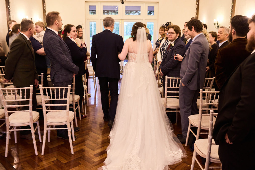 Bride walking down the aisle, seen from behind, as guests stand for the wedding ceremony at Crondon Park