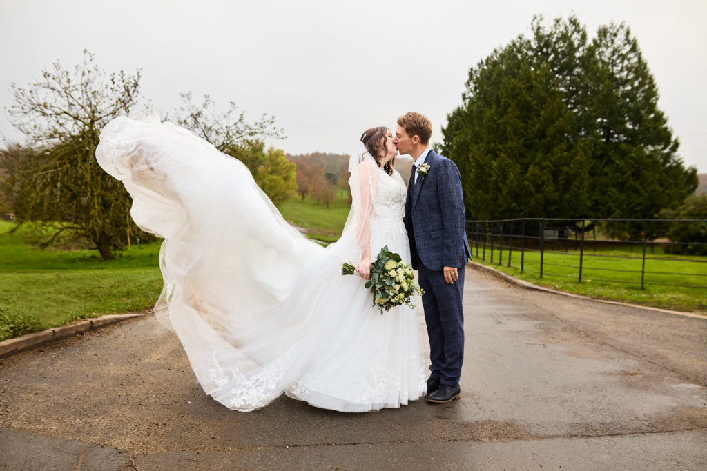 Bride and groom kissing on the driveway at Crondon Park as her dress train billows in the wind