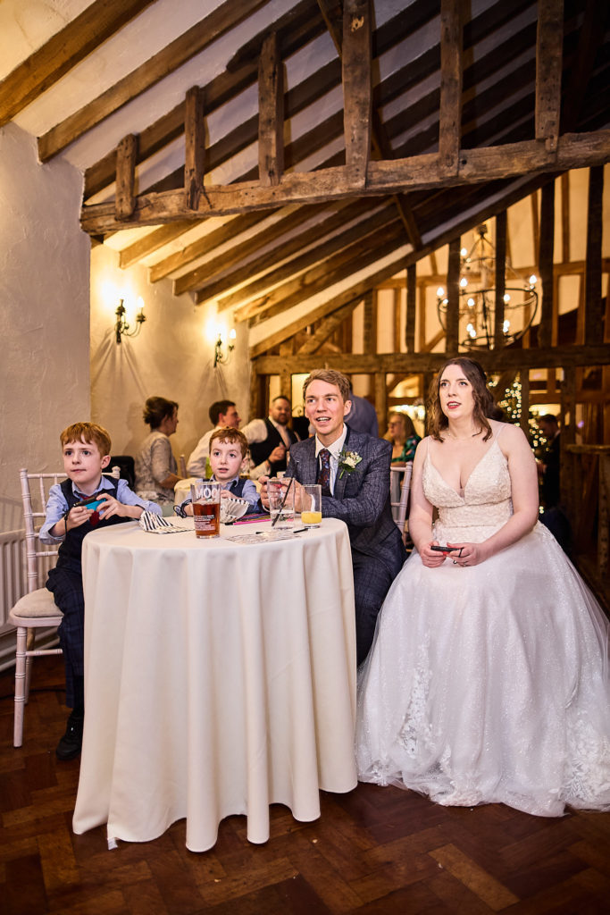 Bride and groom sitting with young guests at a round table in the oak-beamed barn