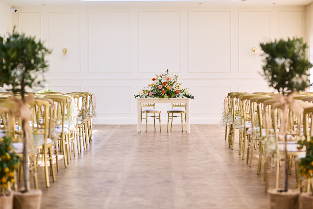 Ceremony room set with gold chairs, olive trees and a flower-topped table at Prested Hall