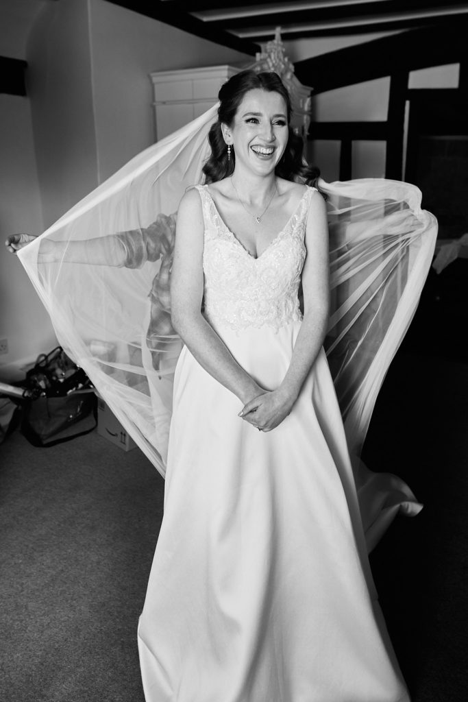 Black and white photo of a bride laughing in her wedding dress as her veil is held out behind her