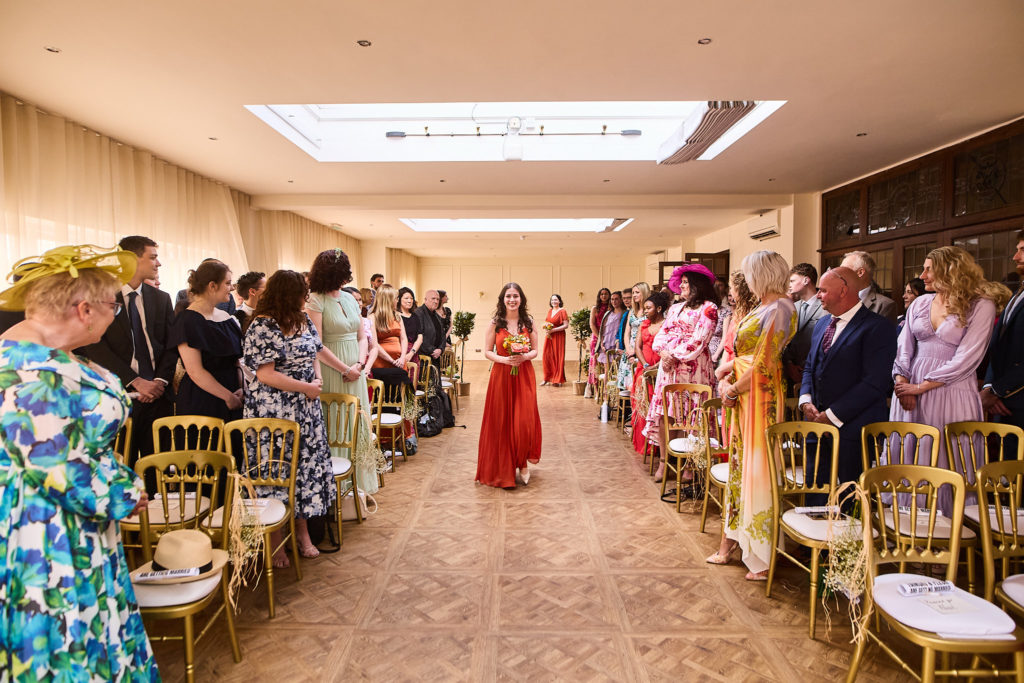 Bridesmaid in a red dress walking down the aisle between guests in colourful outfits