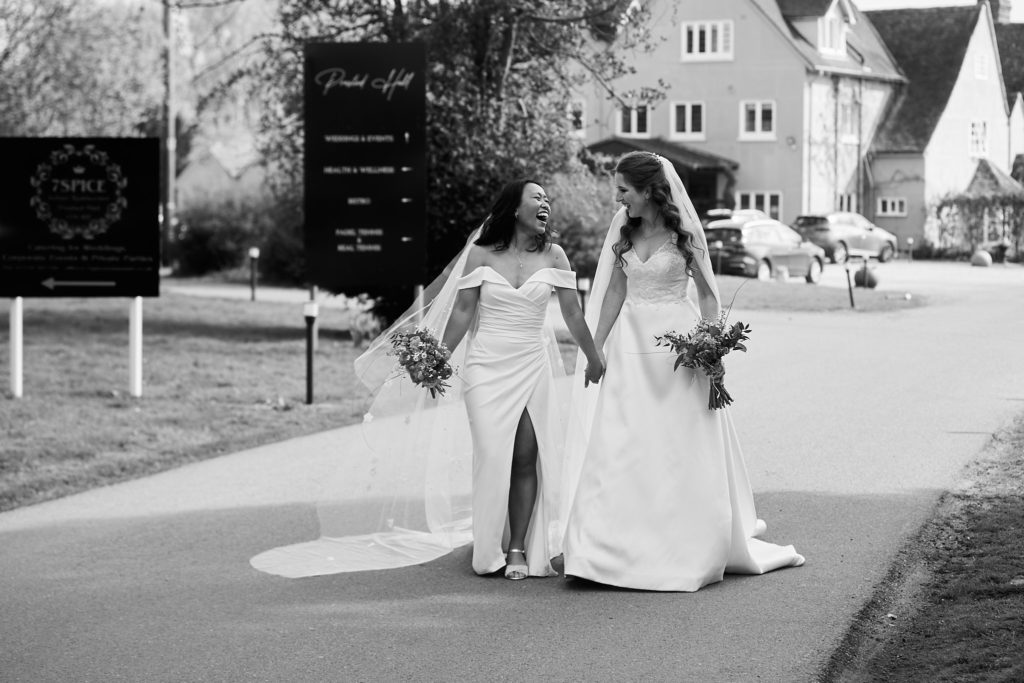 Black and white photo of the two brides walking arm in arm along the drive at Prested Hall
