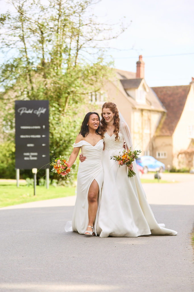 Two brides laughing as they walk arm in arm past the Prested Hall sign on their LGBTQ wedding day