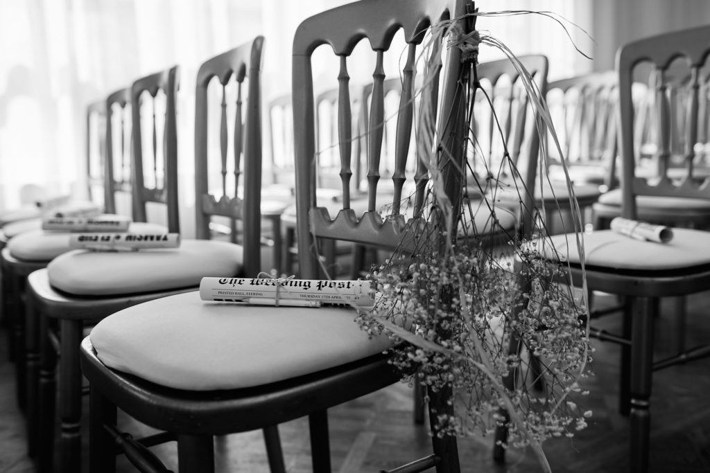 Black and white photo of rows of ceremony chairs with a wedding newspaper on each seat