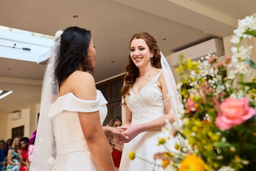 Two brides smiling at each other at the start of their LGBTQ wedding ceremony at Prested Hall