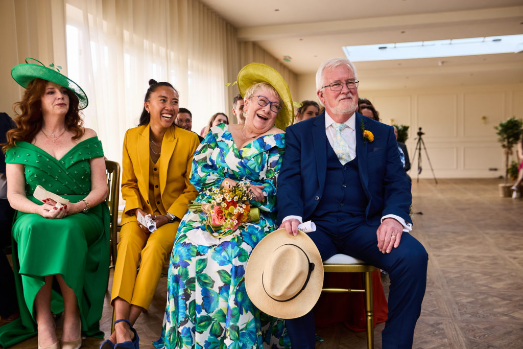 Front-row guests in brightly coloured outfits laughing during the ceremony