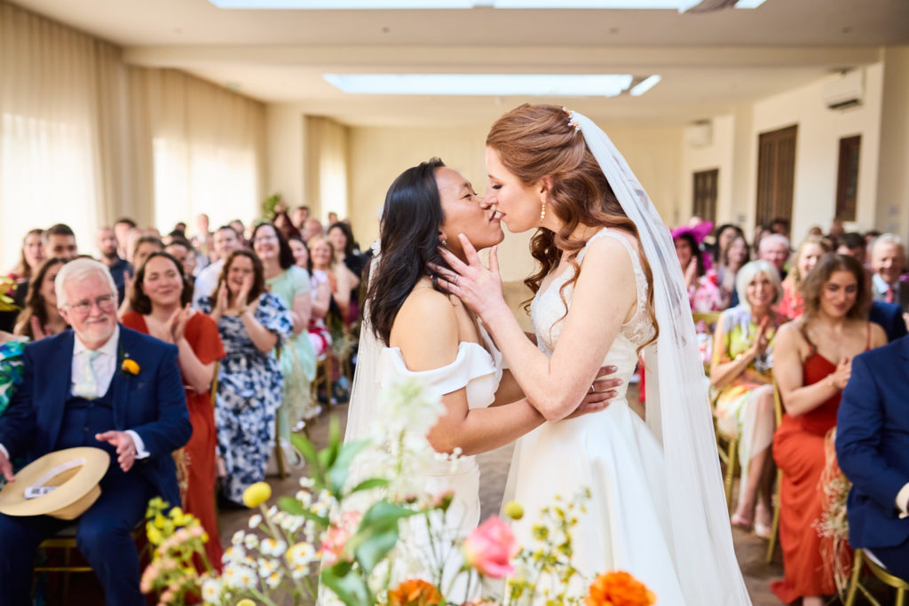 Two brides sharing their first kiss as guests cheer at their same-sex wedding at Prested Hall