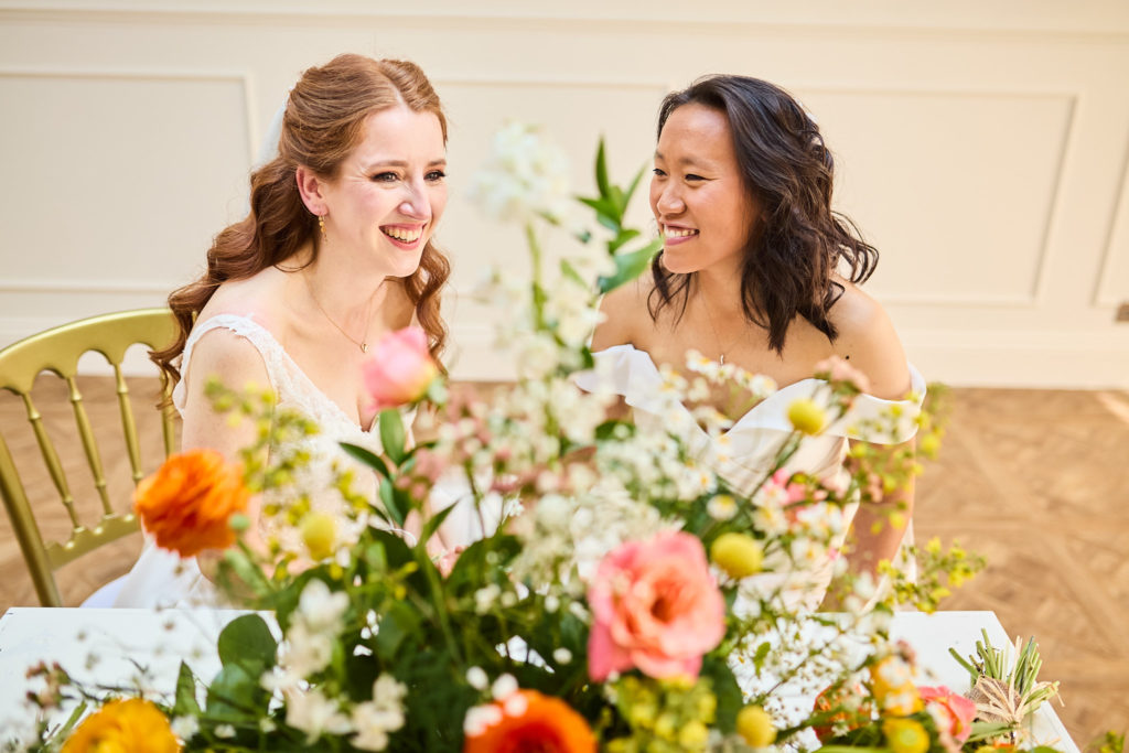 Two brides laughing as they sign the register behind a display of flowers