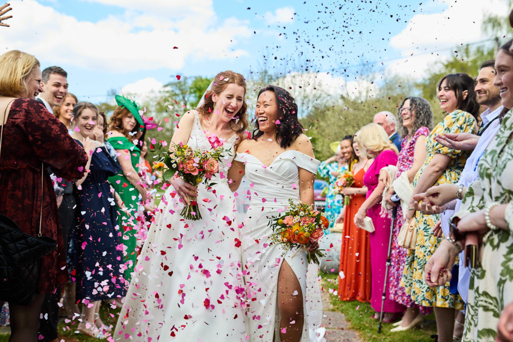 Two brides walking through a shower of confetti at their LGBTQ wedding at Prested Hall, Colchester