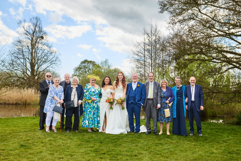 Two brides with their families beside the lake at Prested Hall