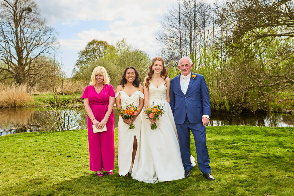 Two brides with a couple in a pink dress and navy suit beside the lake
