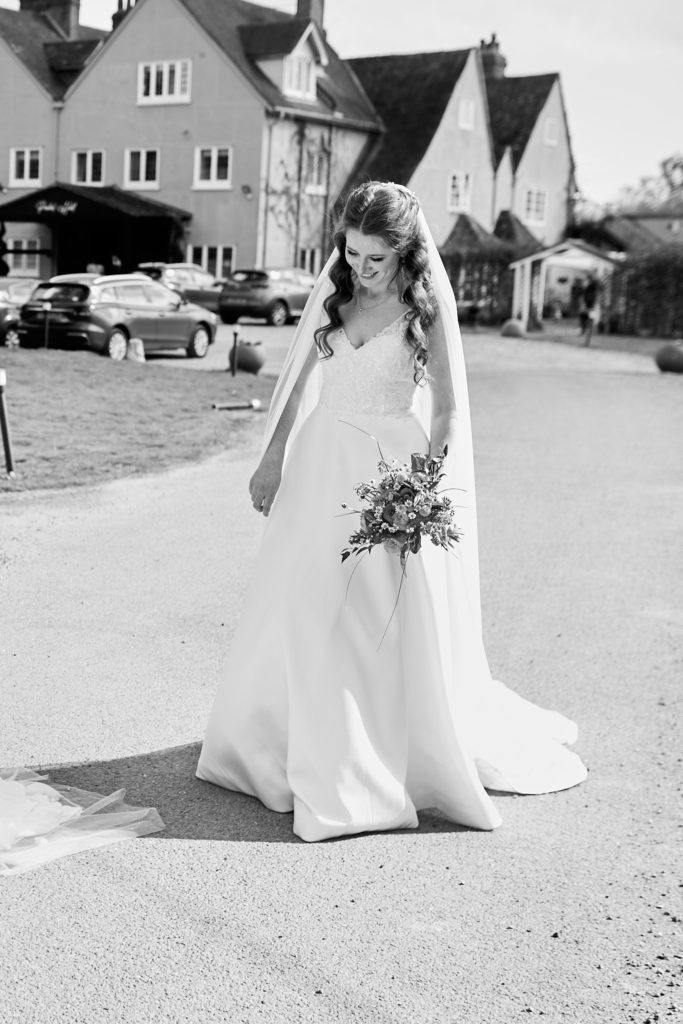 Black and white photo of a bride walking along the drive at Prested Hall with her bouquet