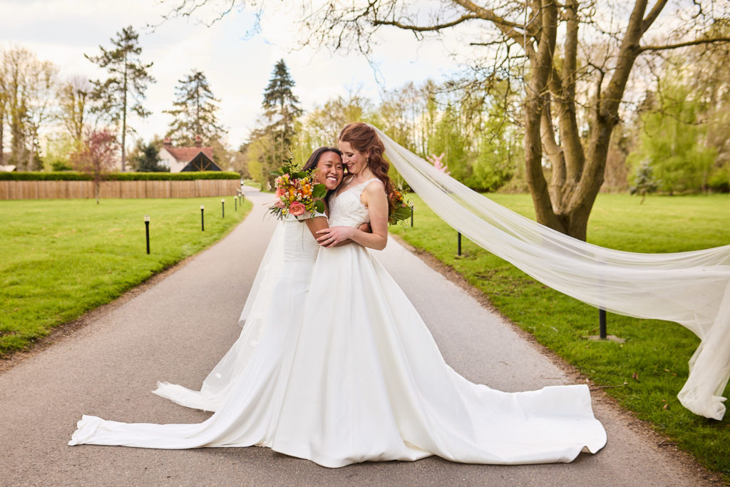 Two brides embracing on the drive at Prested Hall as their veils blow out behind them