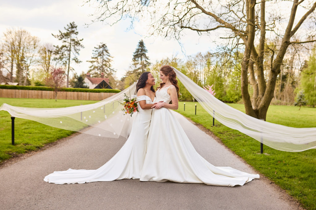 Two brides face to face on the tree-lined drive at Prested Hall, veils streaming in the wind at their same-sex wedding