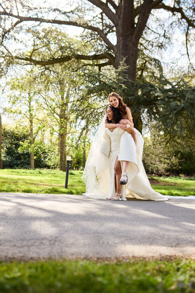Bride giving her wife a piggyback under the trees at Prested Hall