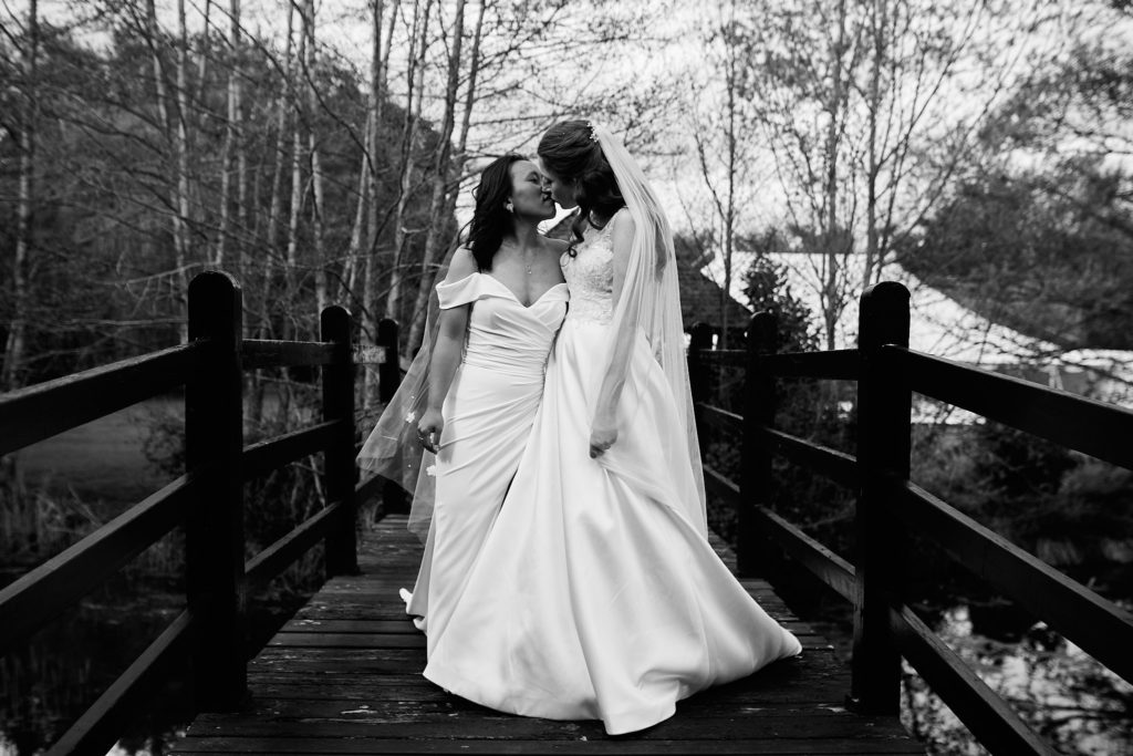Black and white photo of the two brides kissing on a wooden bridge at their LGBTQ wedding at Prested Hall