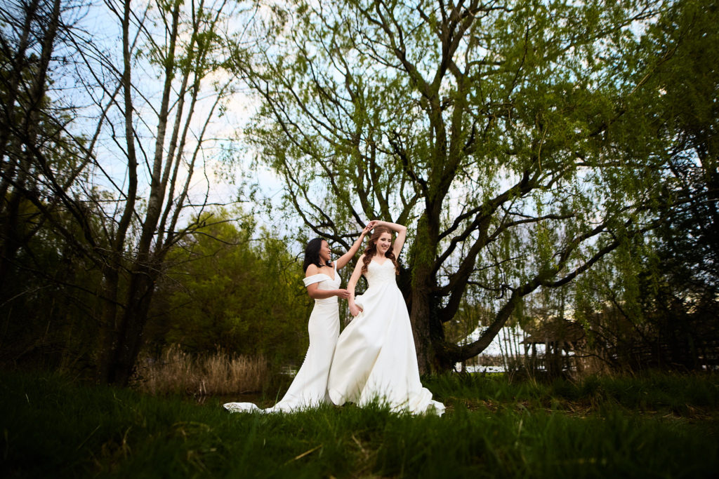 Two brides standing together beneath willow trees at Prested Hall