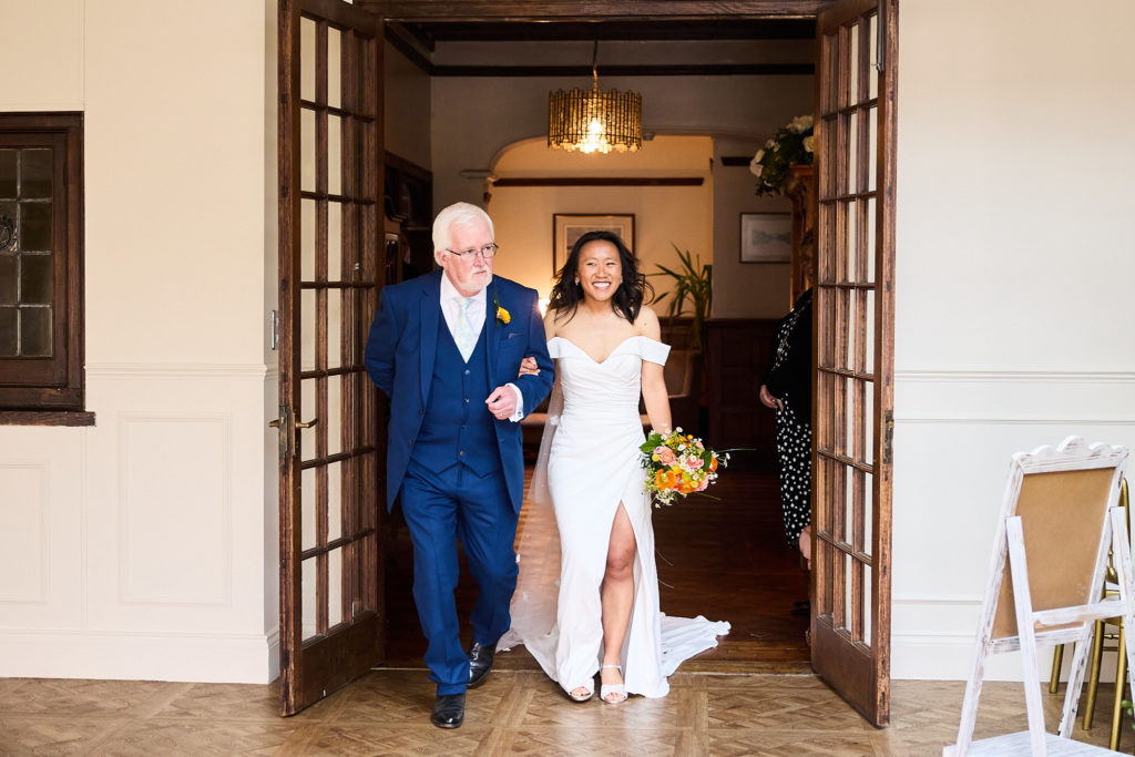 Bride arriving through double doors on the arm of an older man in a navy suit
