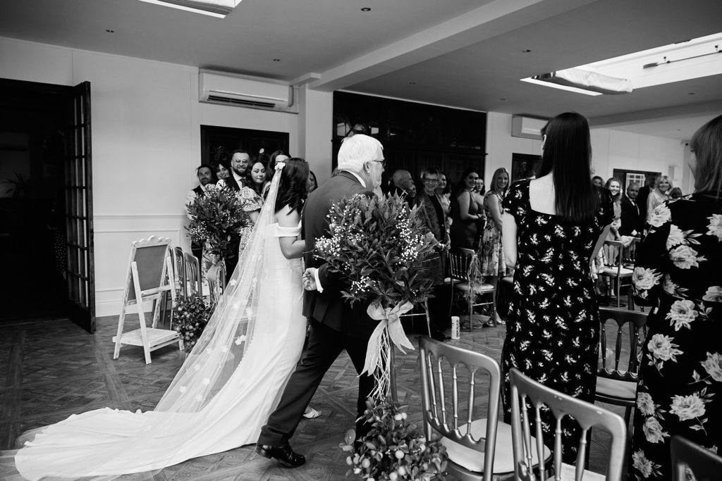 Black and white photo of a bride walking down the aisle with her long veil trailing