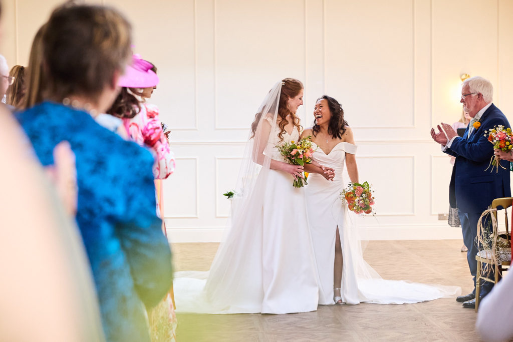 Two brides meeting at the front of the ceremony room as guests look on